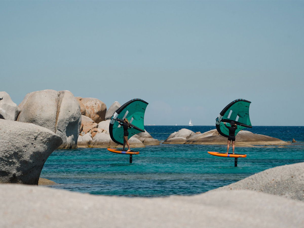 Two windsurfers on a turquoise sea with rocky shoreline.