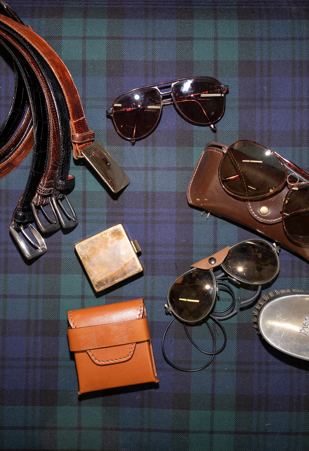 Assorted sunglasses, belts, and leather accessories on a plaid background.