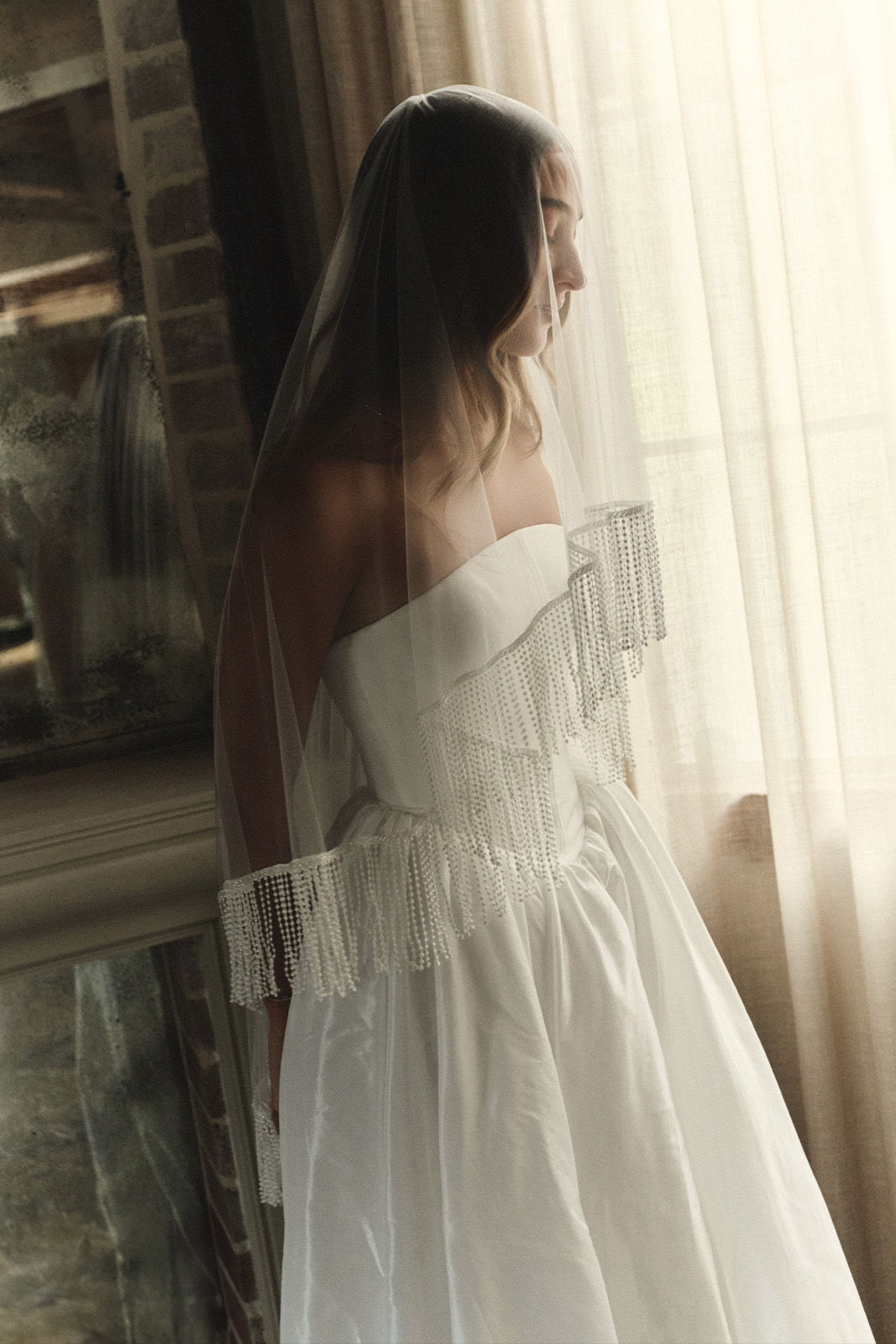 Bride in a white dress and veil, standing by a window.