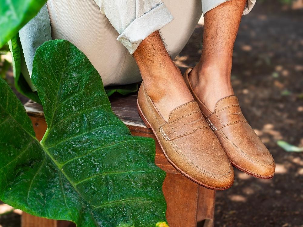 An outdoor lifestyle product shot from the calves down of a person sitting on a wooden bench or box with ankles crossed, wearing light-colored rolled-up trousers. The person is wearing light brown or tan textured leather penny loafers without socks, partially framed by a large, vibrant green elephant ear leaf in the left foreground.