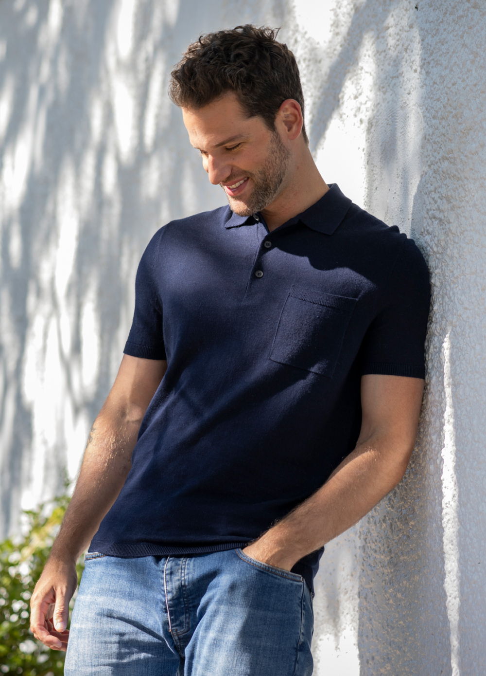 Man wearing a navy polo shirt, leaning against a wall, looking down.