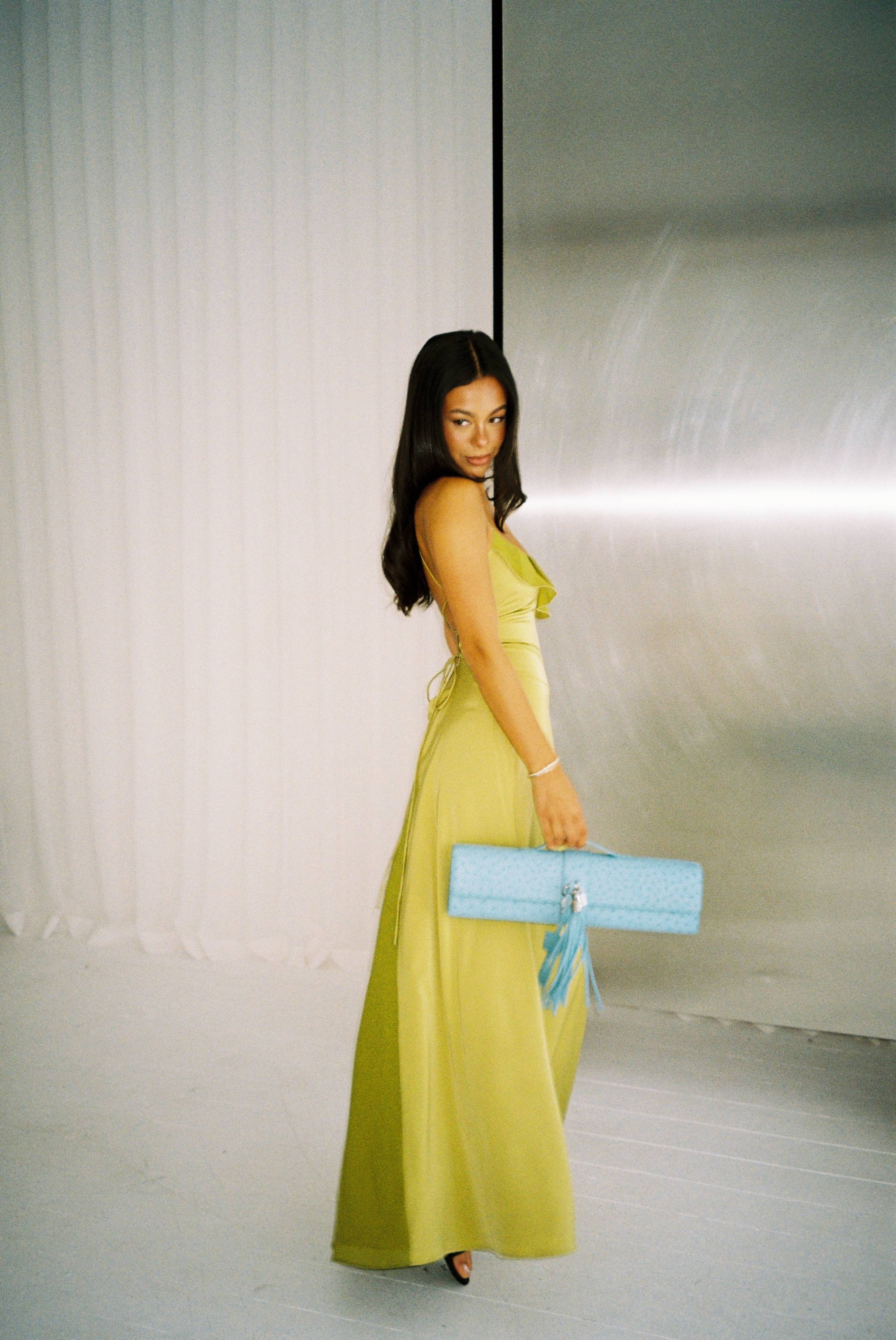 Woman in a yellow dress holding a blue bag, posing in a studio setting.