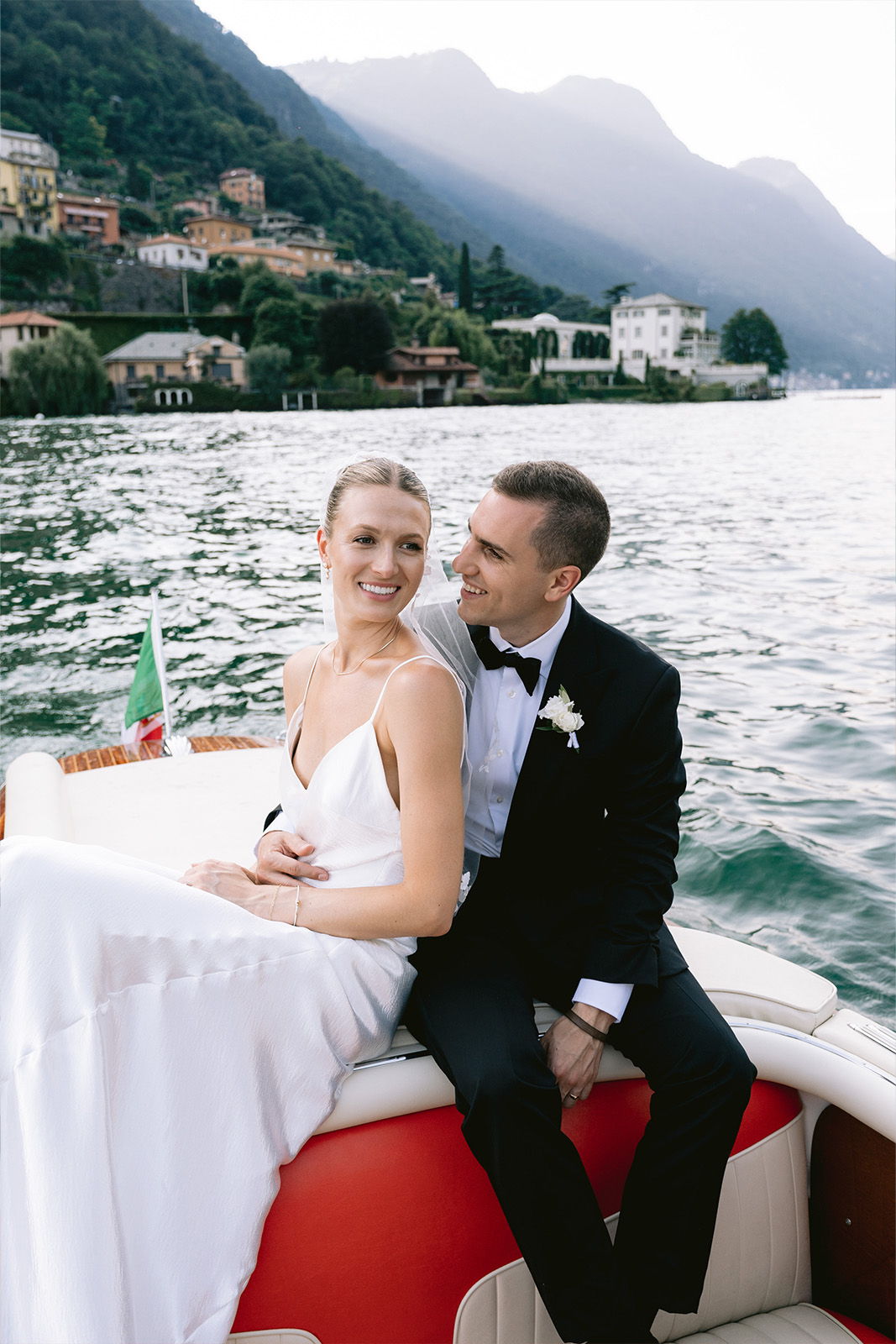 Bride and groom sitting on a boat, smiling by a lake.