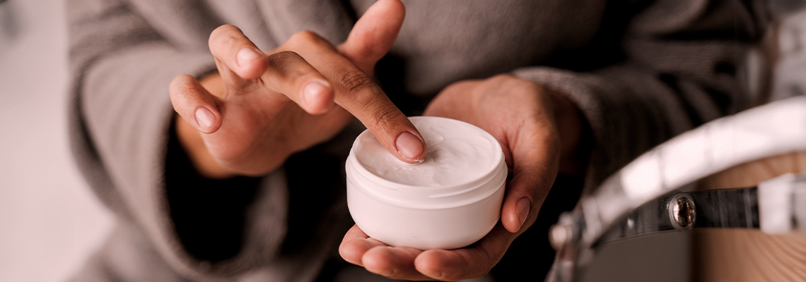 Person applying cream from a jar while wearing a robe.