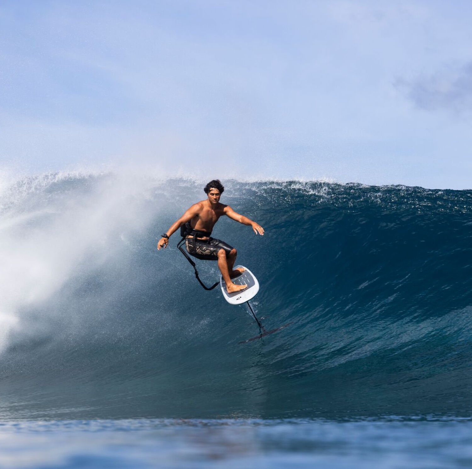 Surfer riding a large wave under a blue sky.