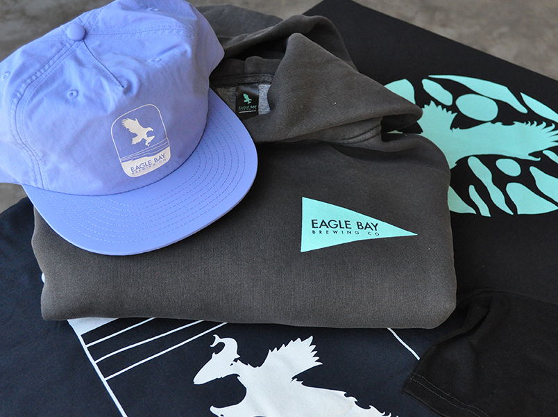 Light blue surf cap and folded grey hoodie with Eagle Bay Brewing Co. teal logo, sitting on screen printed tees with eagle bay brewing co designs