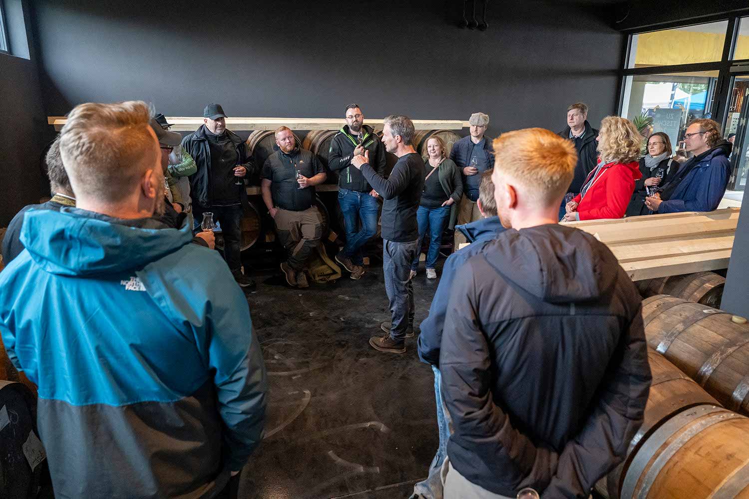 Group of people gathered indoors, listening to a speaker near wooden barrels.