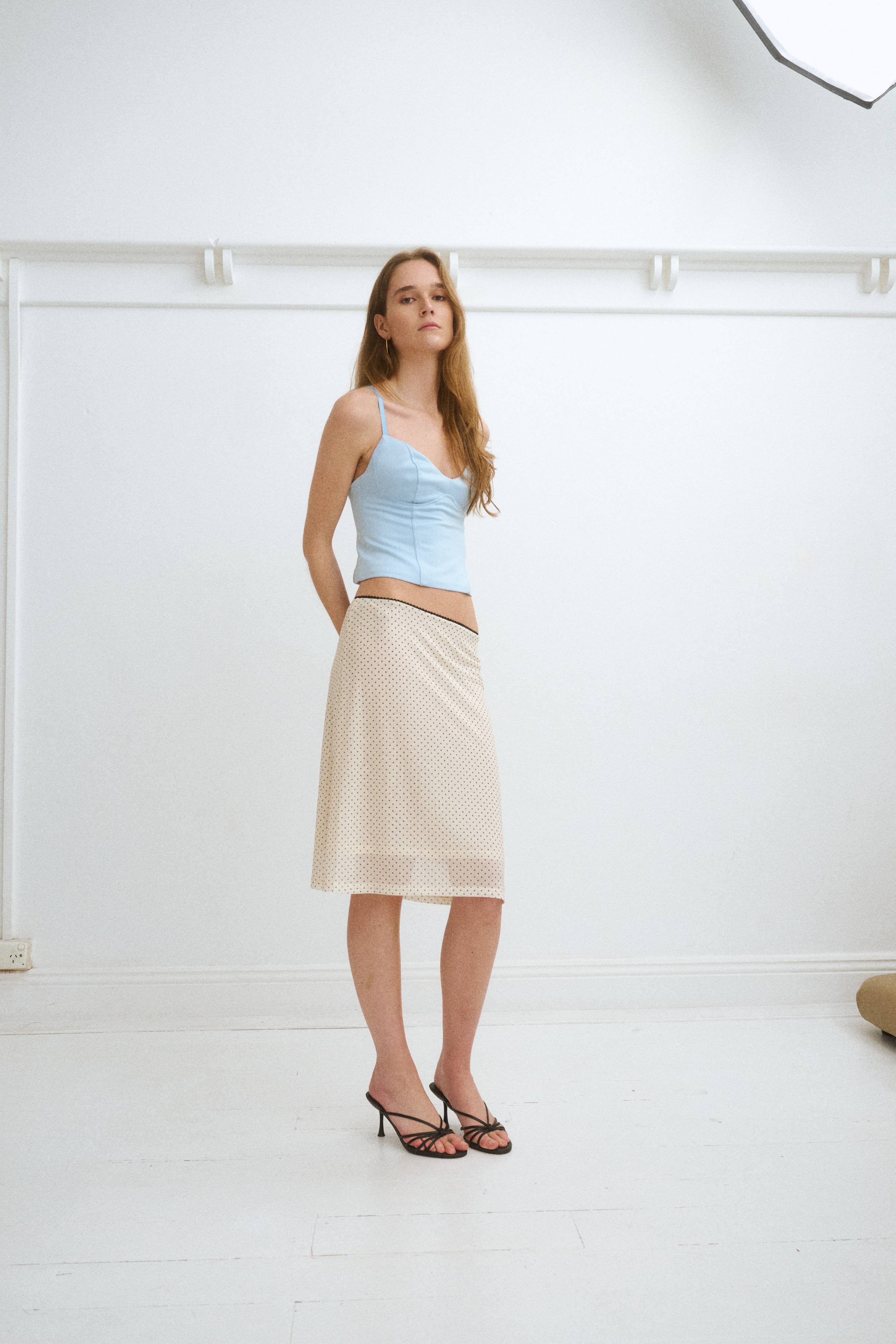 Model in a light blue top and beige skirt, standing in a studio setting.