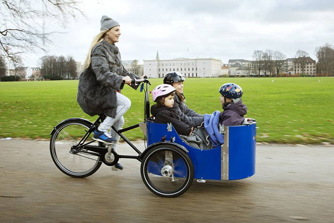 A mother rides a Nihola 4.0 cargo bike in a public park with three children in the front box.
