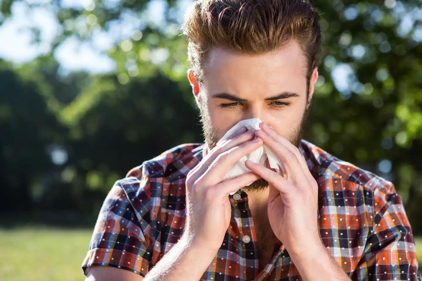 Man met pollenallergie, oftewel hooikoorts niest in een zakdoekje. Hij is omgeven door gras en bomen waar veel pollen in de lucht zitten. Niezen, tranende ogen zijn symptomen van allergie.