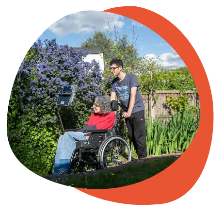 Person in wheelchair outdoors, assisted by another individual, surrounded by flowers.