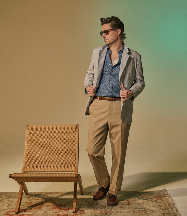 A stylish individual in a suit poses beside a chair against a neutral background.