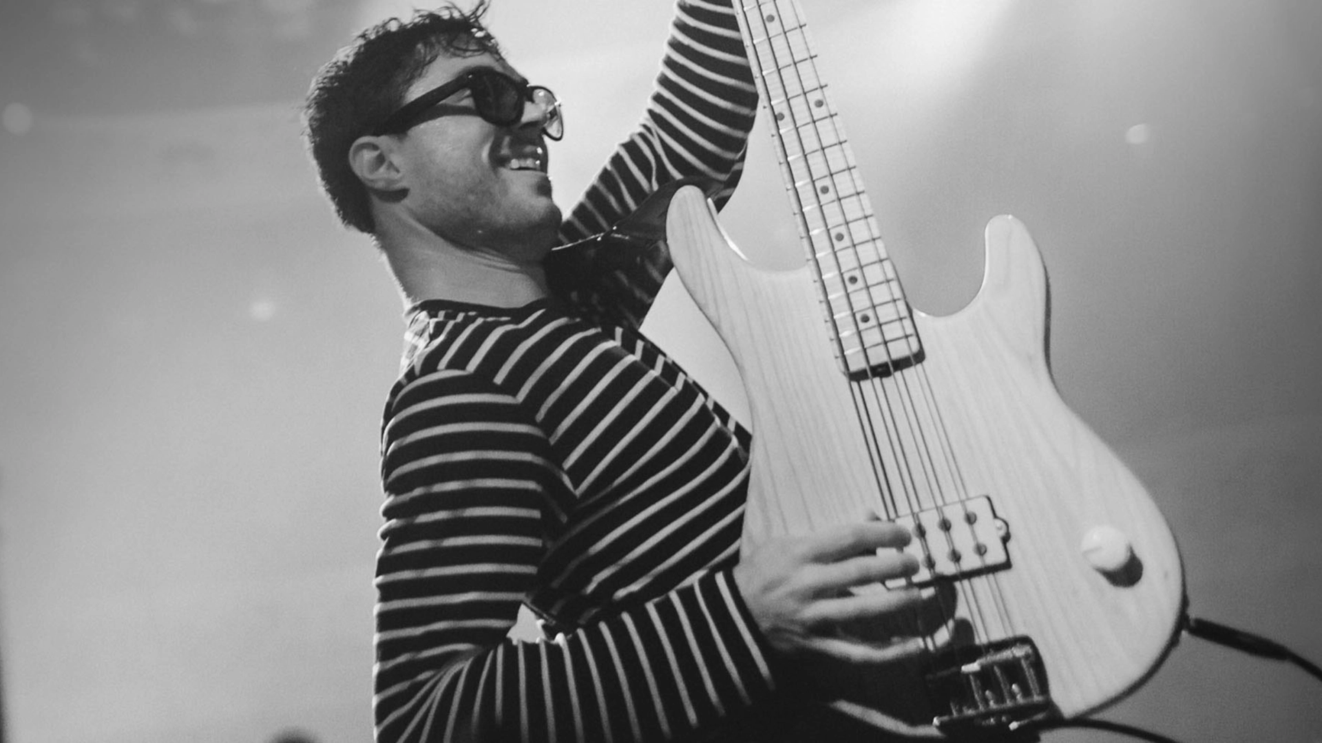 Joe Dart playing his Music Man signature bass guitar on stage, wearing sunglasses and a striped shirt.