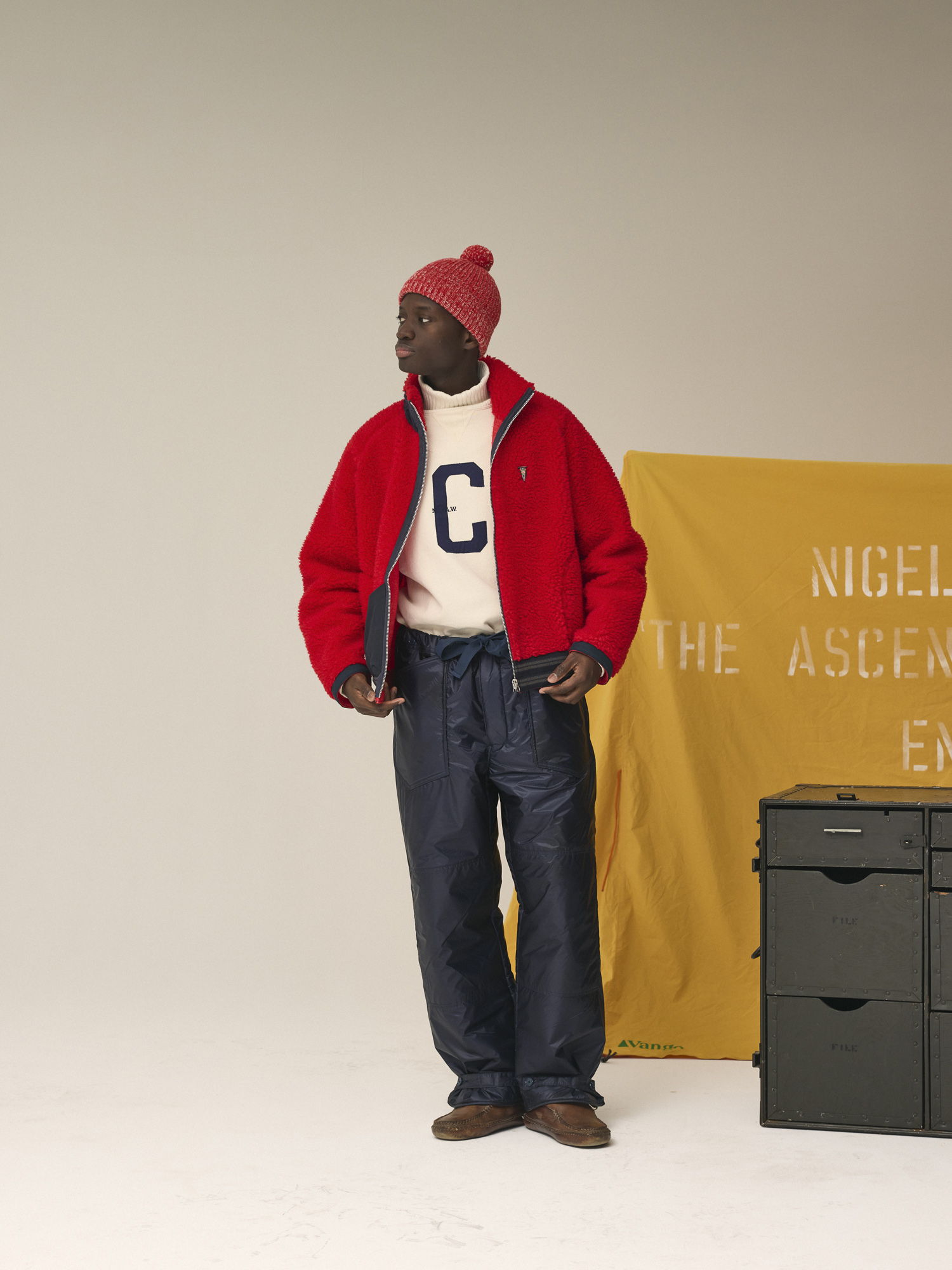 Person wearing red jacket and hat, standing beside yellow backdrop and storage boxes.