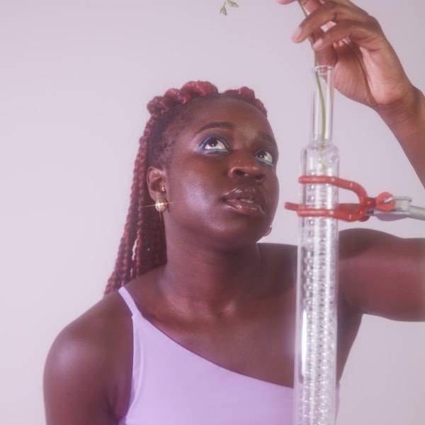Fotografia de mulher olhando para cima, segurando o ramo de uma planta que sai de um tubo de vidro à sua frente