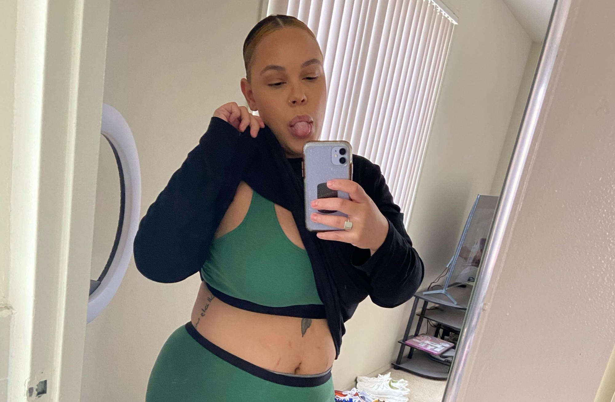 A woman pulls up her black sweater to expose her green bralette while she takes a mirror selfie with her tongue sticking out.