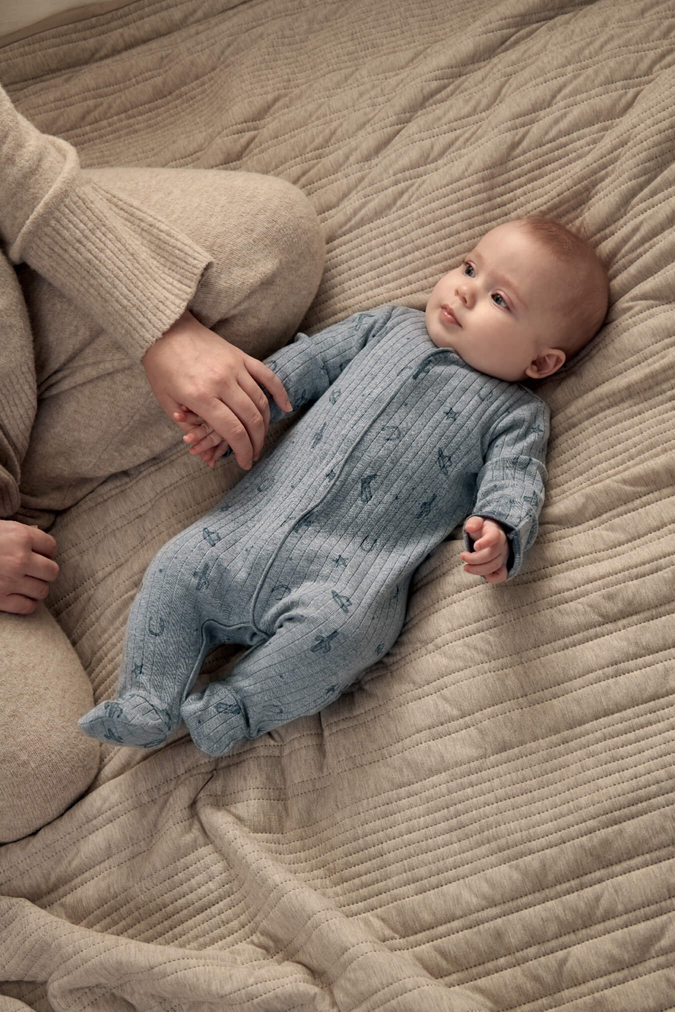 Baby in a blue onesie lying on a textured blanket, holding an adult's hand.
