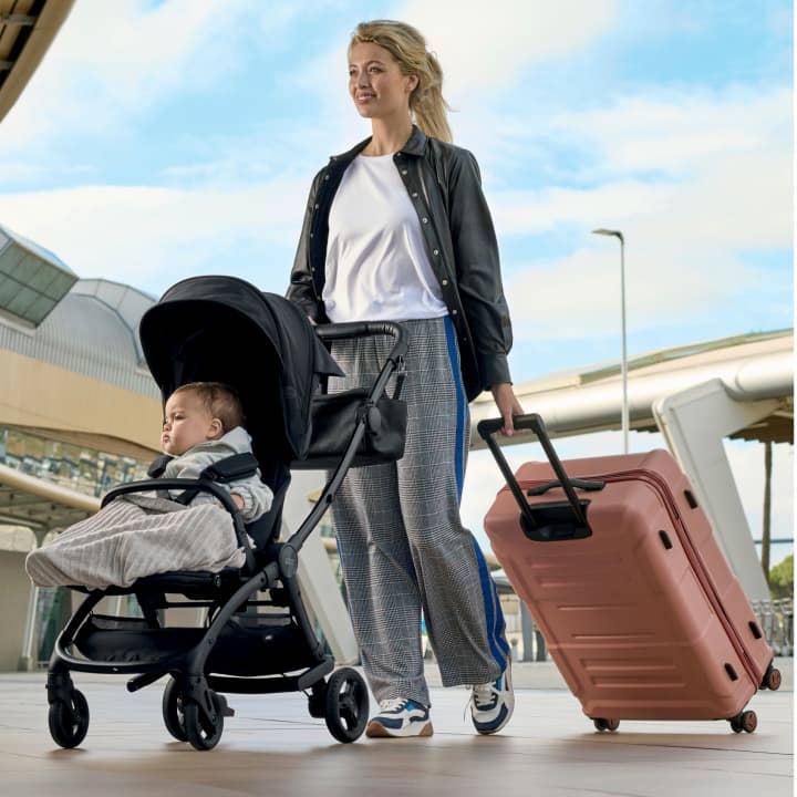 A mother is outside the airport pushing her child in the black Airo pushchair and pulling a suitcase behind her.