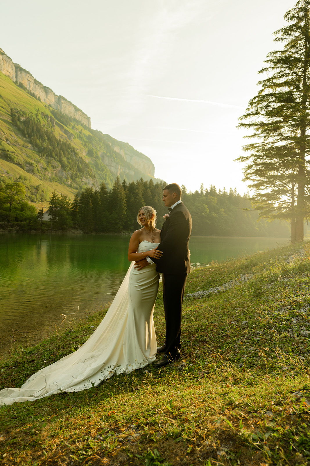 Bride and groom standing by a serene lake at sunset.