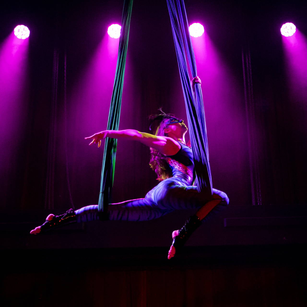 Aerialist performing on silks, wearing a blue mask and leotard on purple backlit background