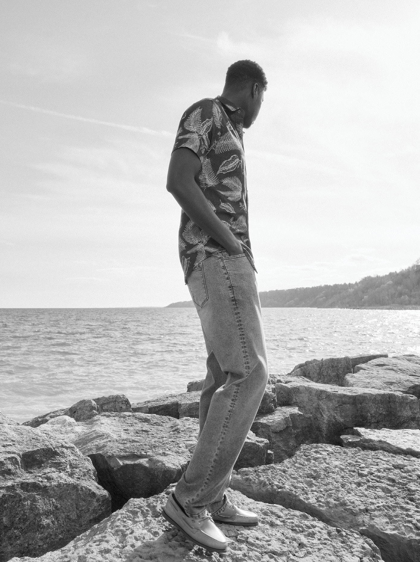Tall man standing on rocks by the water wearing a floral shirt and light wash jeans