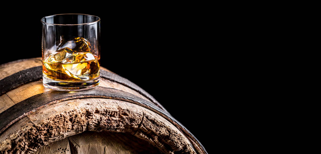 A Glass of malt whisky with ice cubes sitting on a whisky barrel