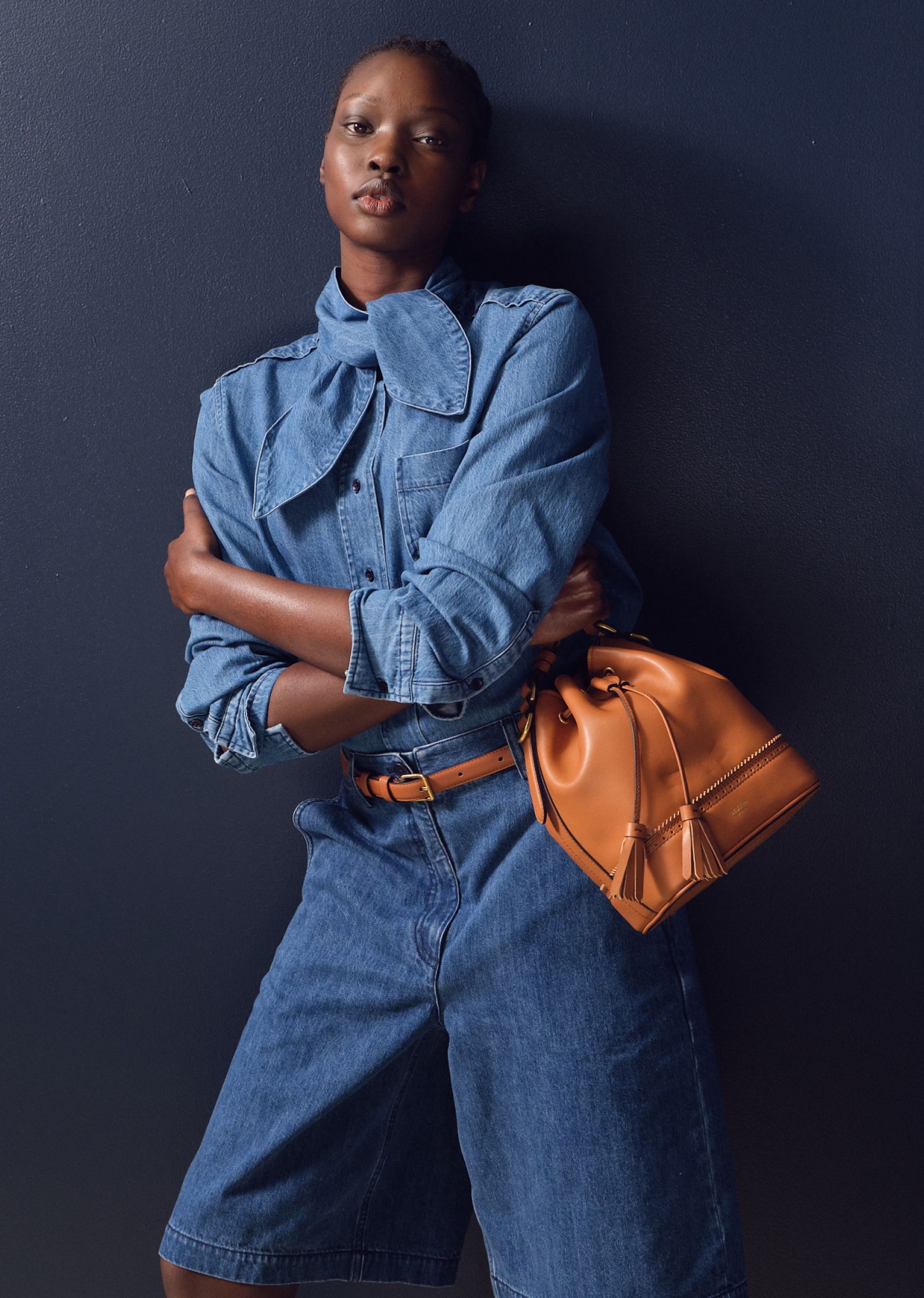 Model in denim outfit holding a brown handbag against a dark background.