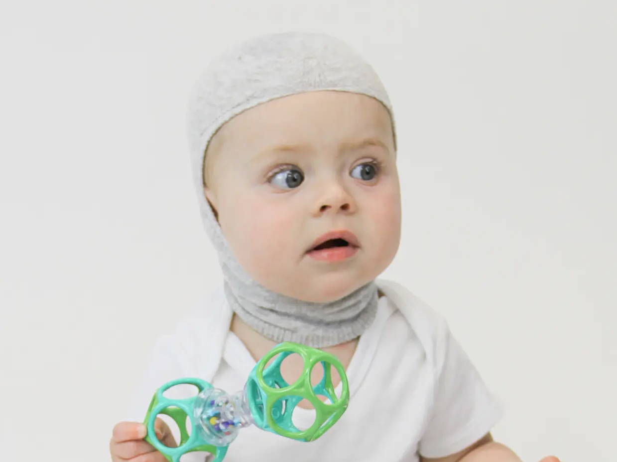 Baby wearing Knit-Rite Balaclava Cranial Interface in grey while holding a toy