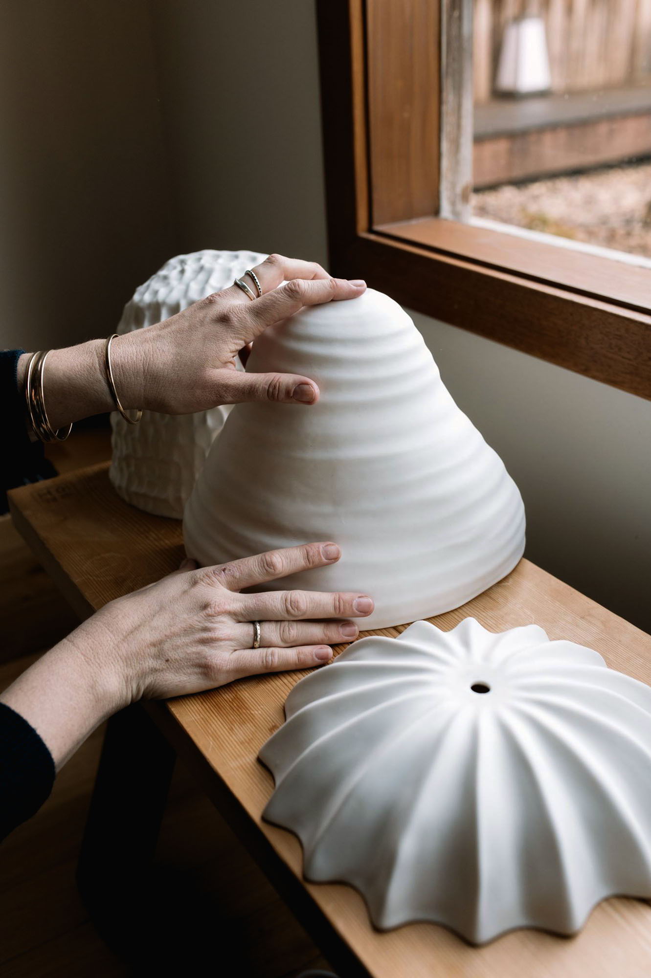 Hands arranging white Milly Dent ceramic wall sconces on a wooden surface near a window.