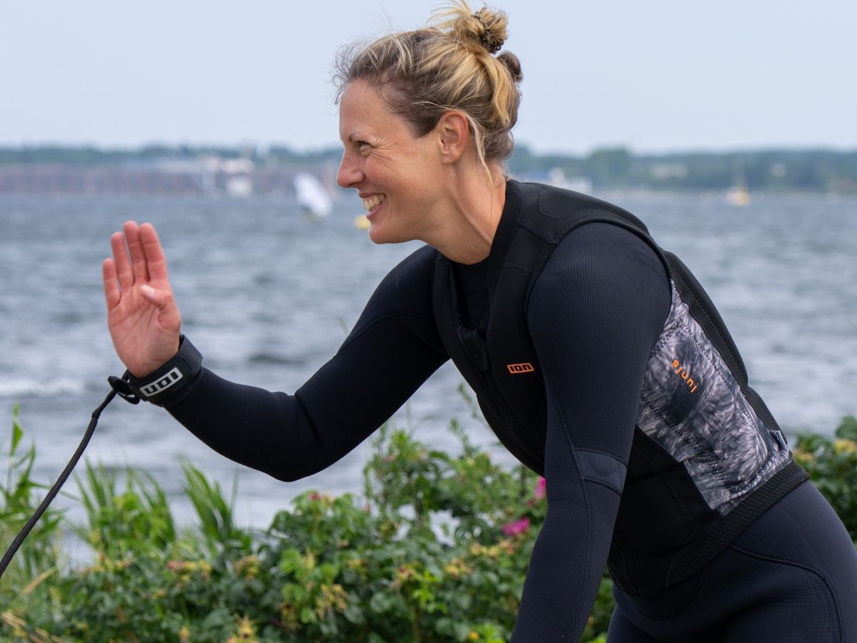 A woman in a wetsuit waves while standing near water.