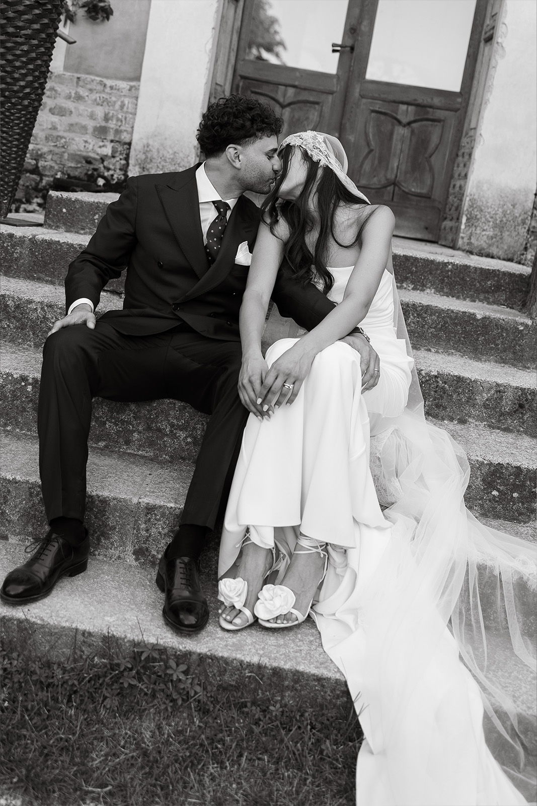 Couple kissing on steps, dressed in wedding attire, surrounded by nature.