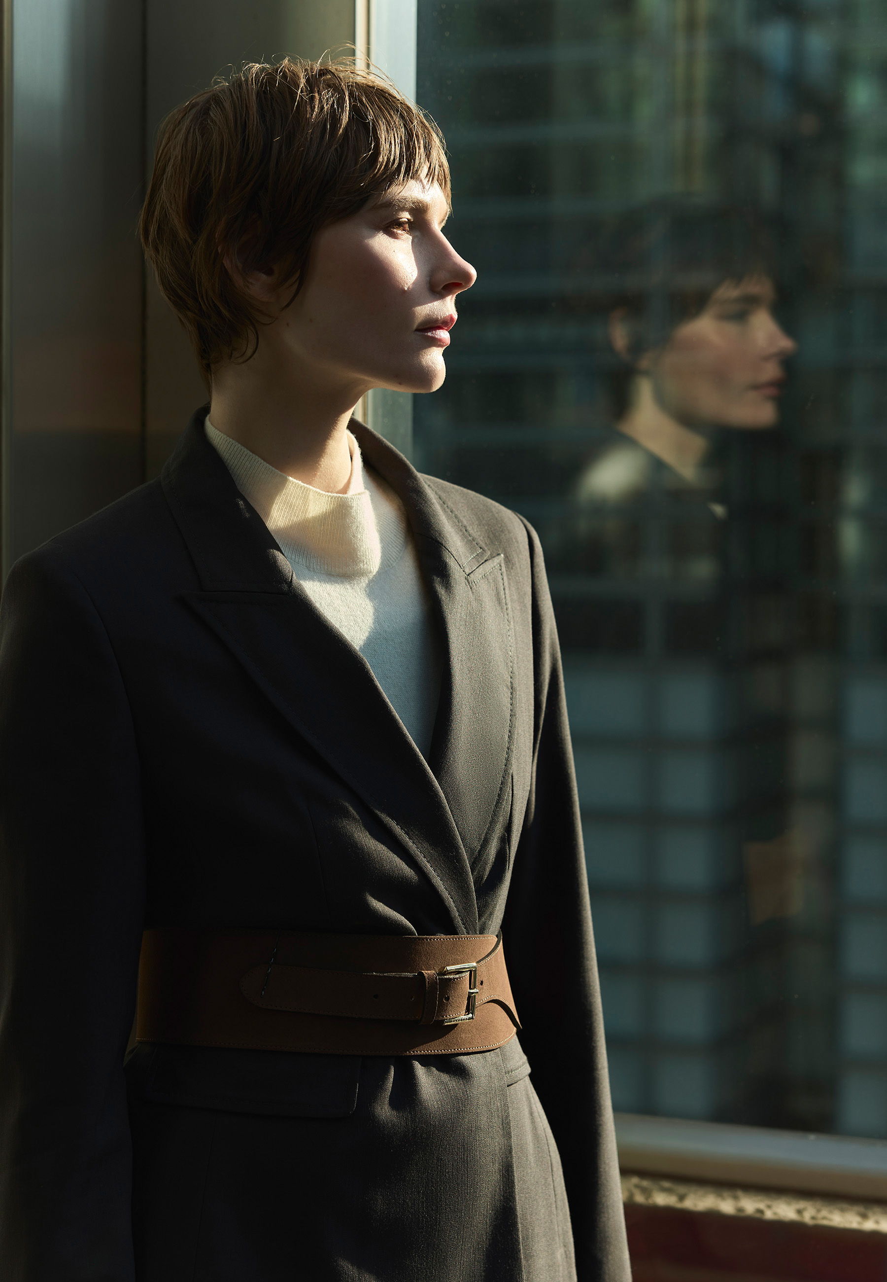 A woman in a suit stands by a window, reflecting cityscape.