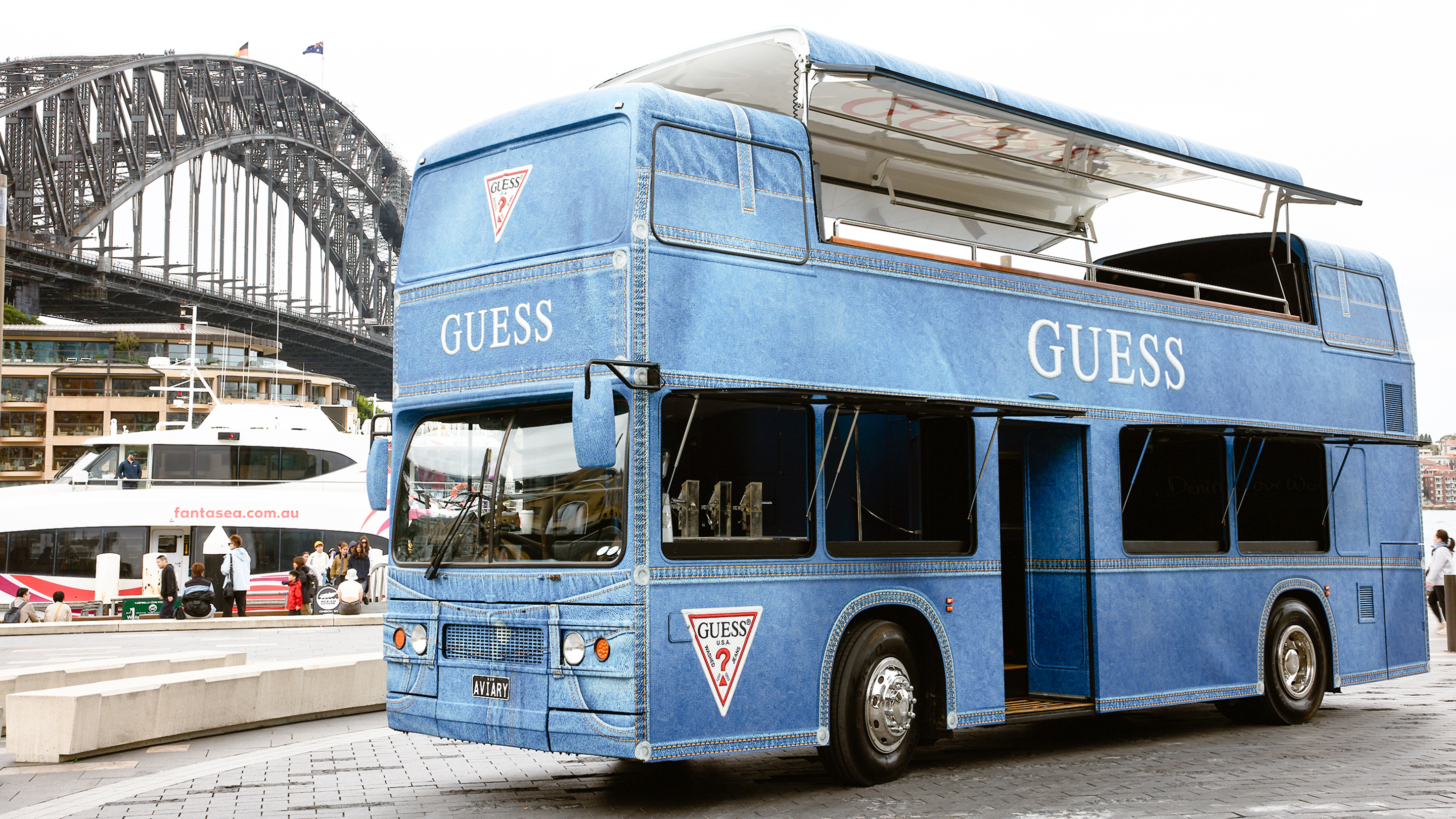 Blue double-decker GUESS denim bus near Sydney Harbour Bridge.