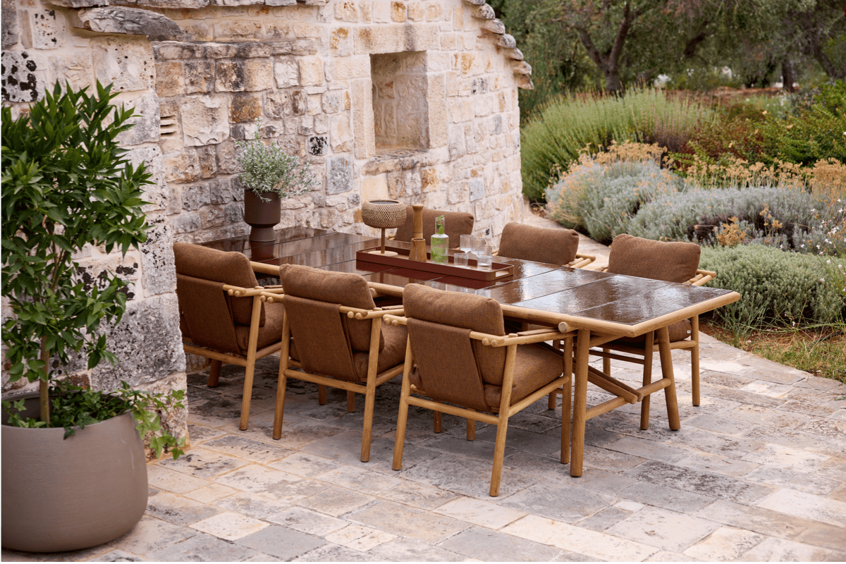 Outdoor dining space with a large rectangular table, brown cushioned dining chairs, and a stone house backdrop surrounded by garden plantings.