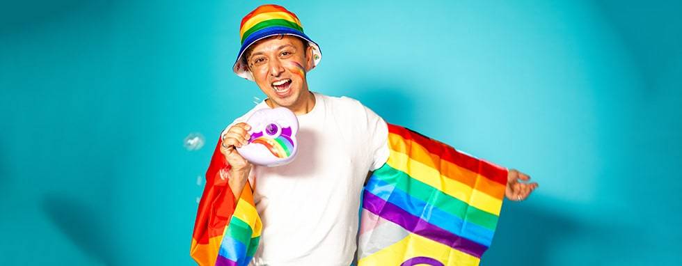 A joyful man wearing a rainbow hat and cape, blowing bubbles from a toy bubble machine against a bright turquoise background.