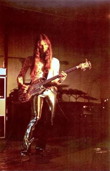 Musician playing bass guitar, wearing shiny pants, with long hair.