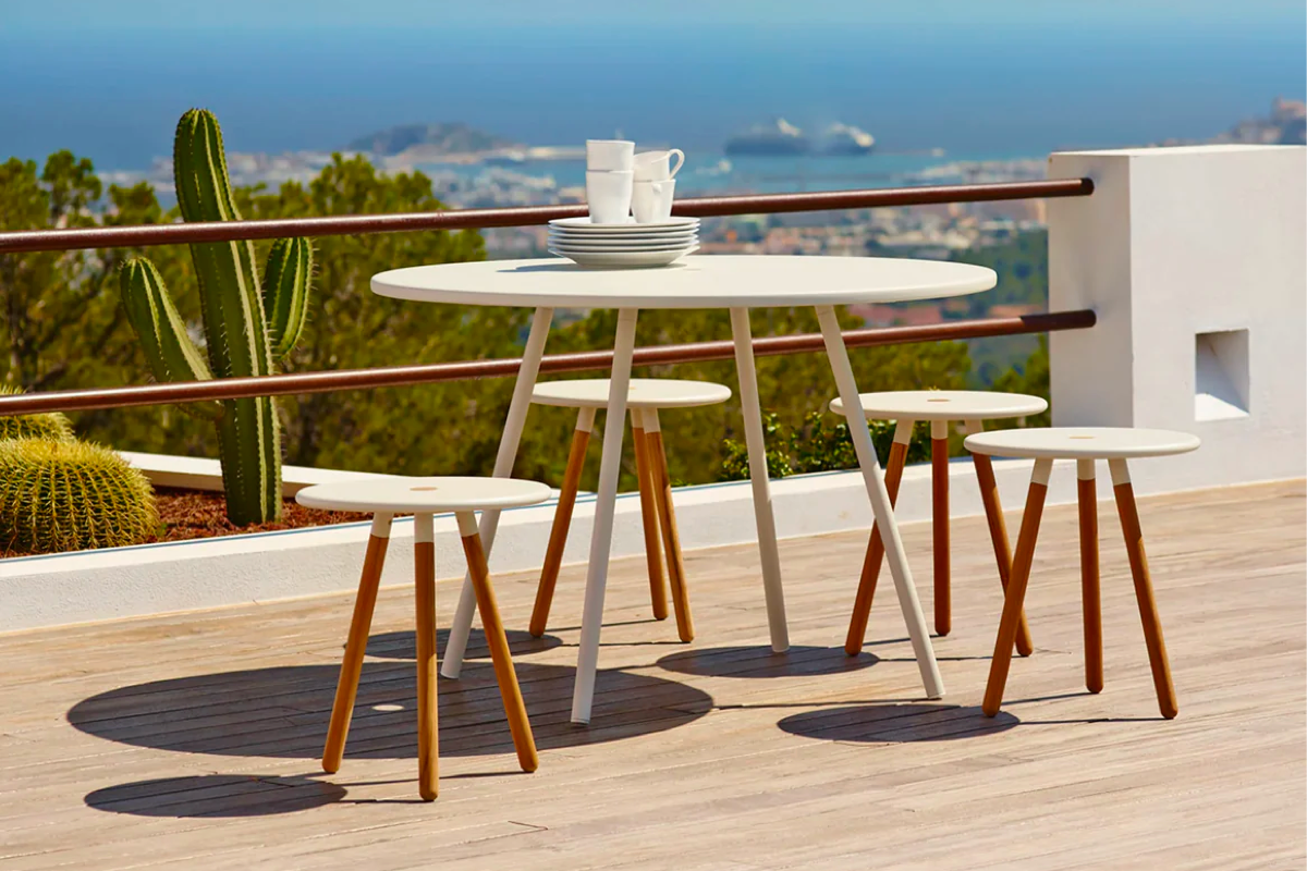 A minimalist Scandinavian style white bistro table and matching stools on a desert balcony with cacti behind.