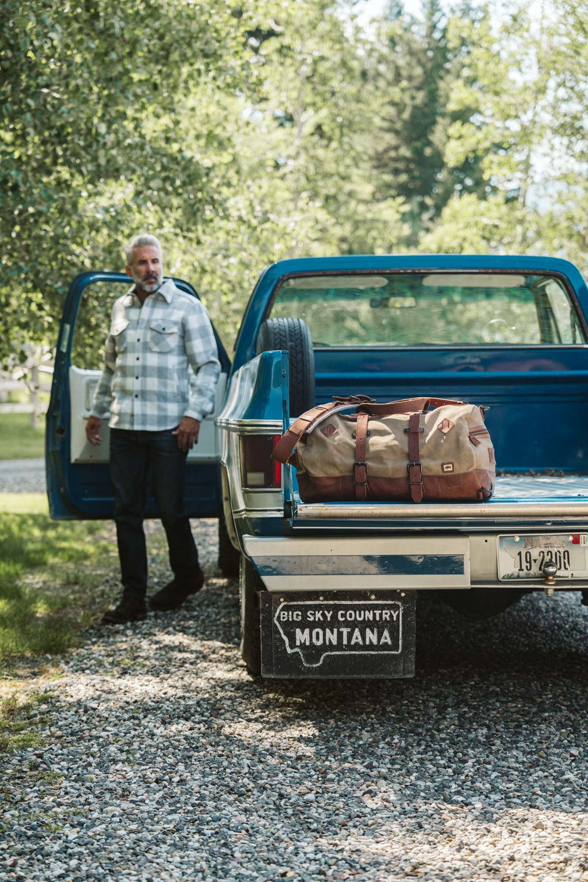 Man beside a vintage blue pickup with a Buffalo Jackson waxed canvas and leather duffle bag in the truck bed