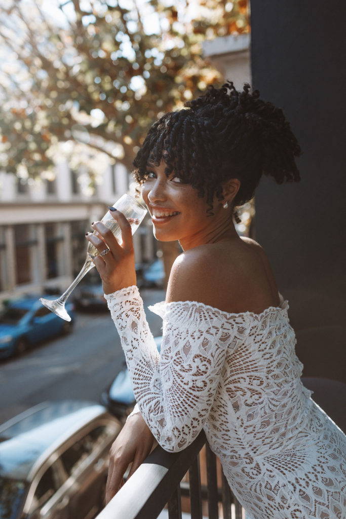 Bride wearing off the shoulder long sleeve lace dress sipping champagne on the balcony