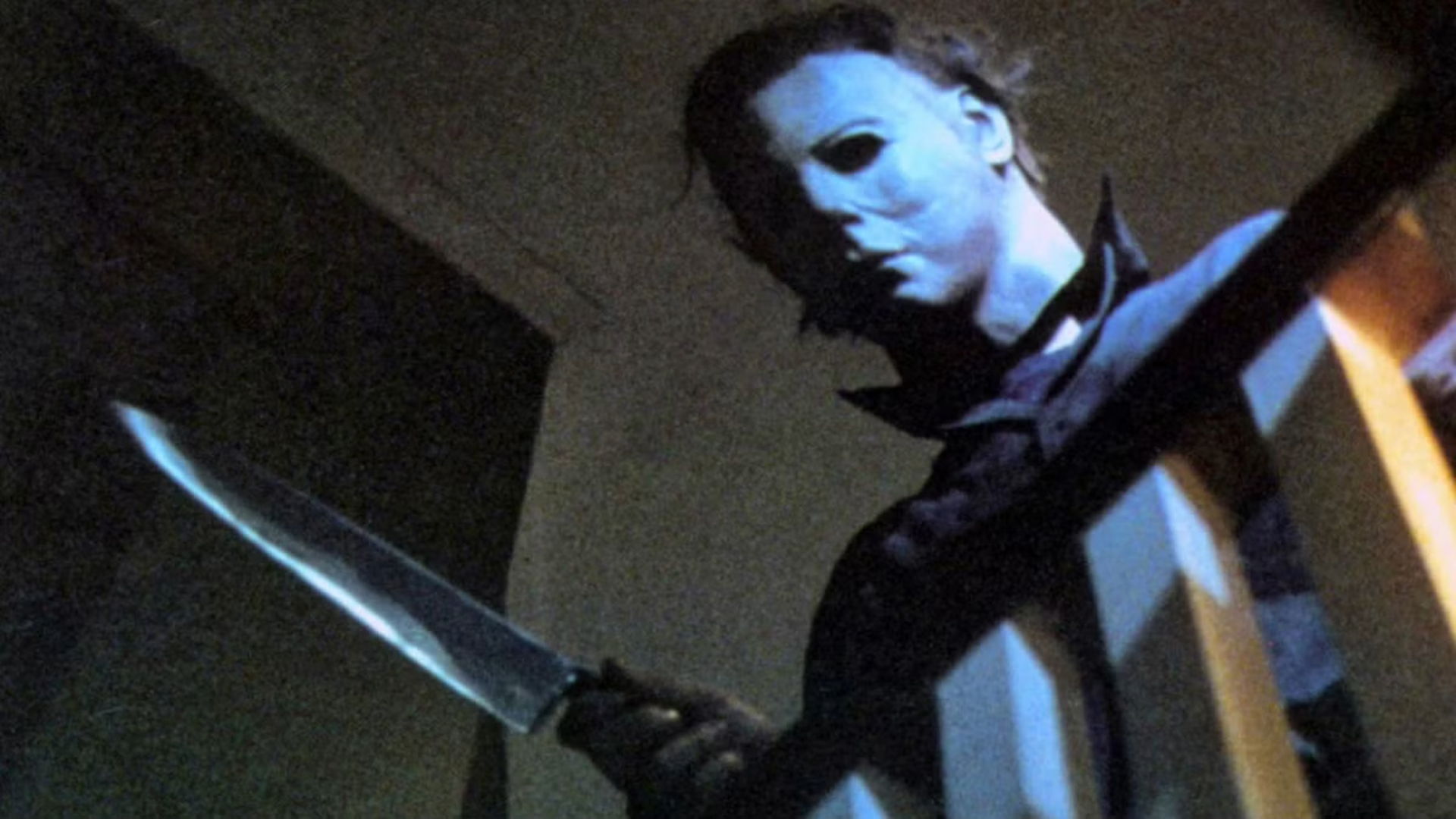 A figure with a mask holding a knife in a dimly lit setting.
