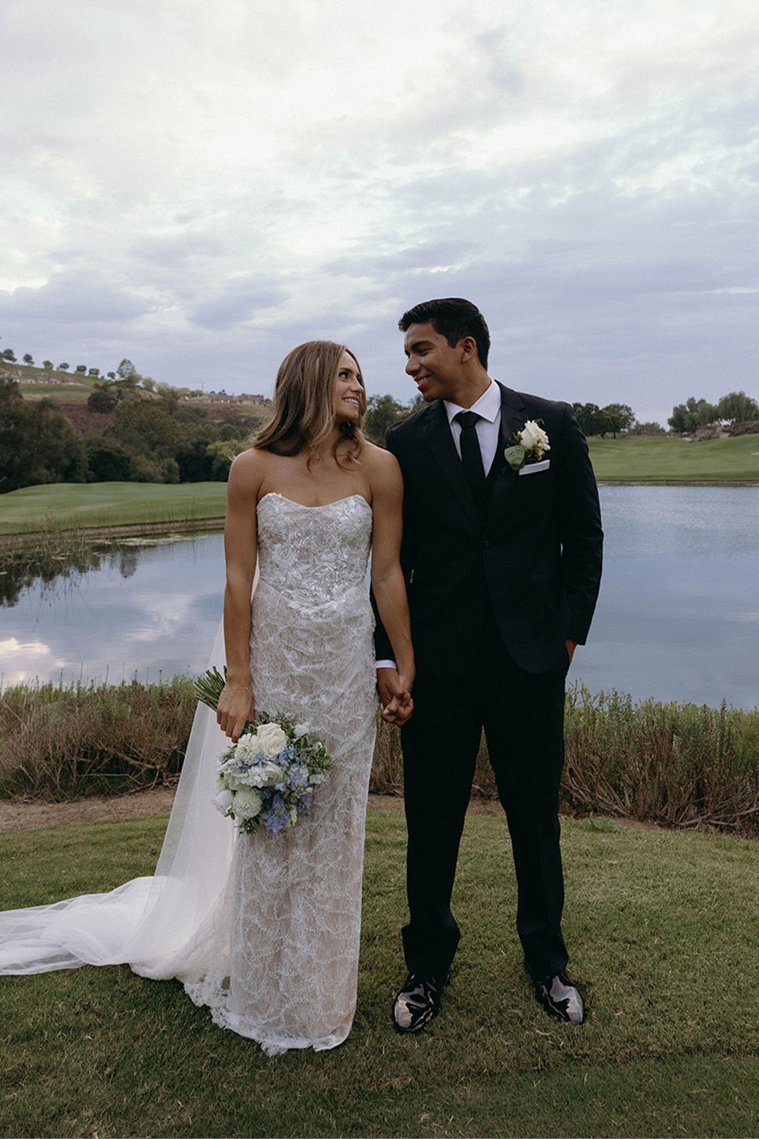 Bride and groom holding hands by a lake, smiling at each other.