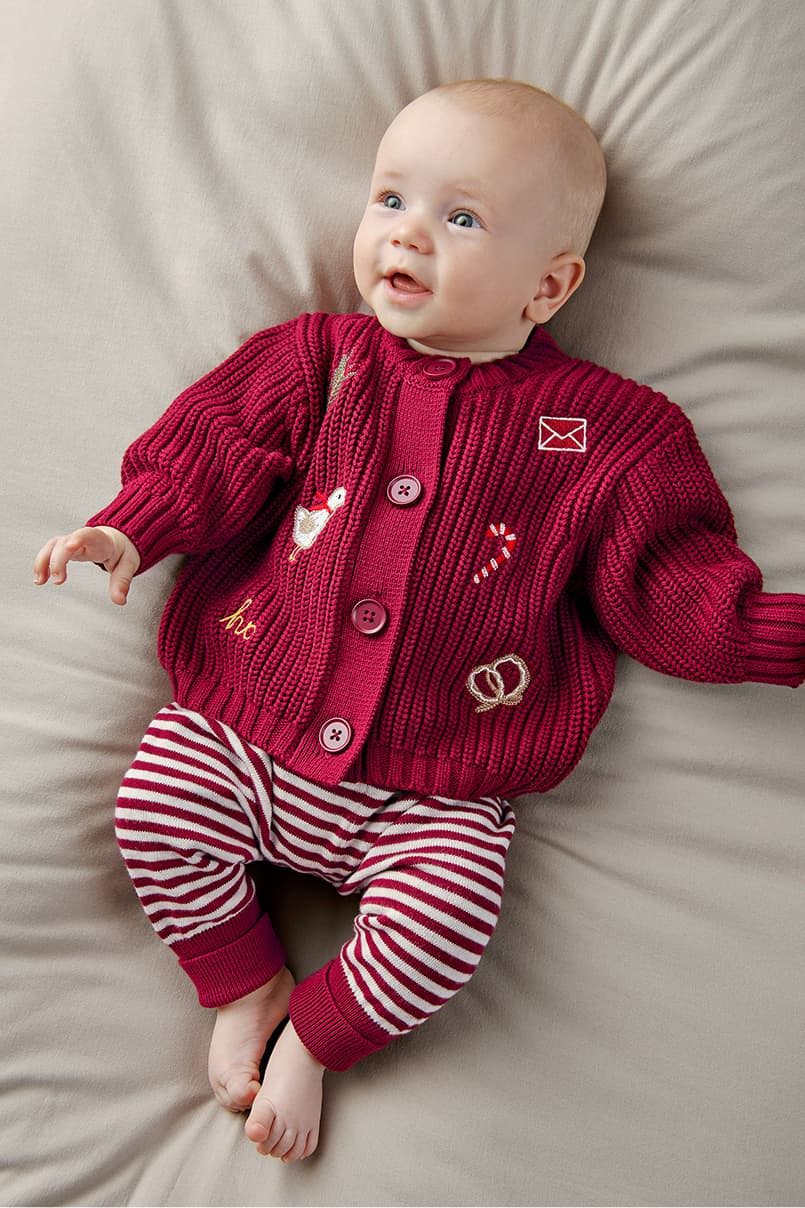 Baby wearing a red cardigan and striped pants, lying on a beige surface.