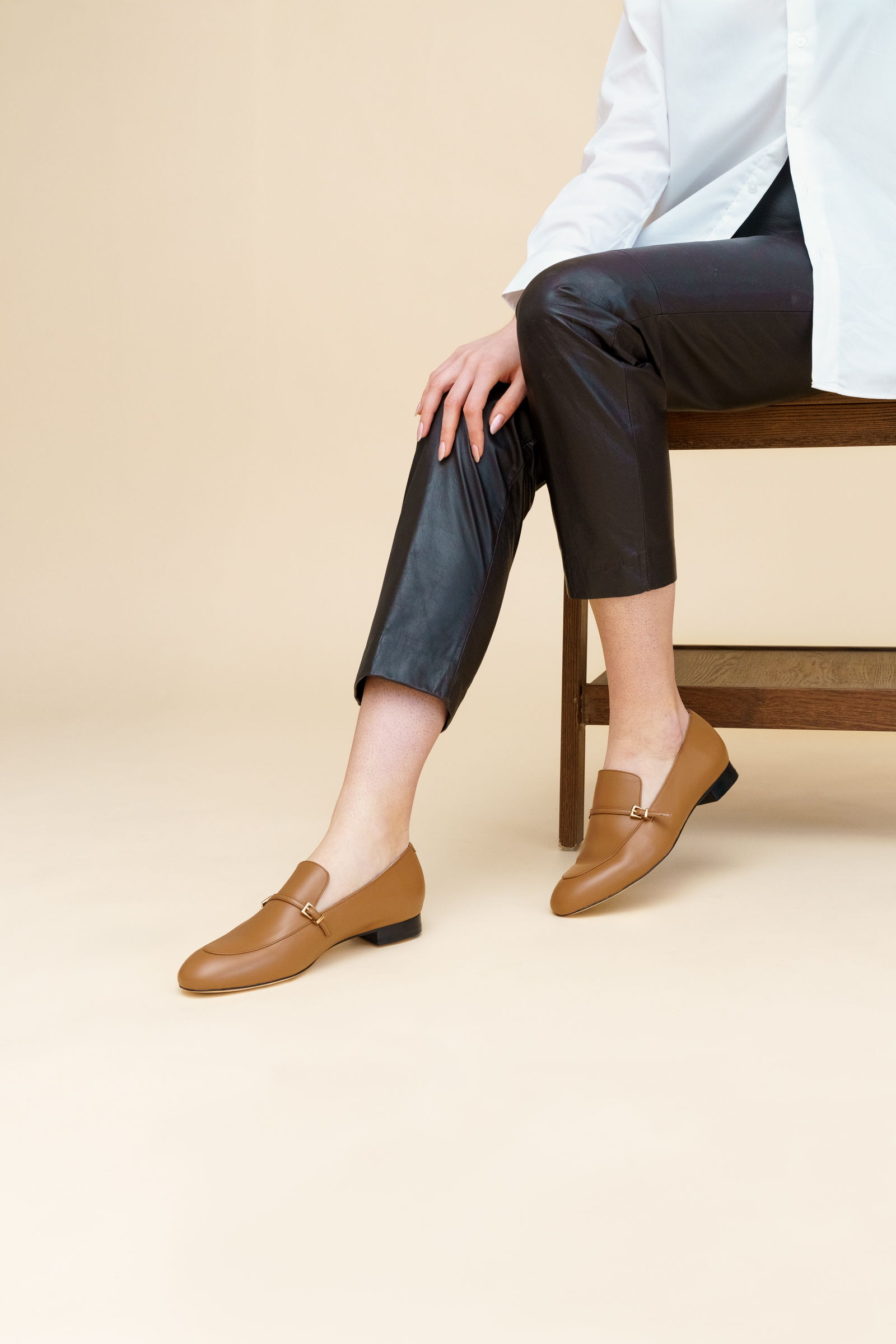 A person wearing brown loafers and black pants sitting on a chair.