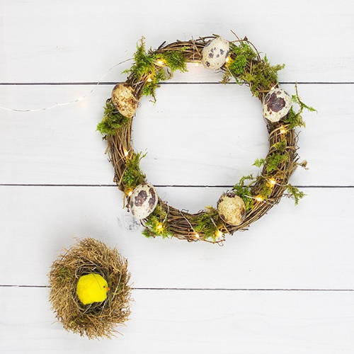 The completed Easter DIY wreath with moss, Easter eggs and micro lights and a small toy chick next to the wreath.