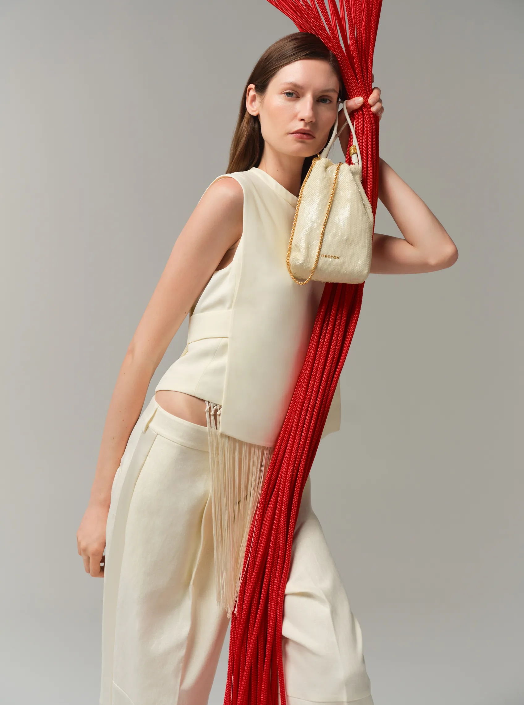 Model in cream outfit holding a bag, with red strands in background.