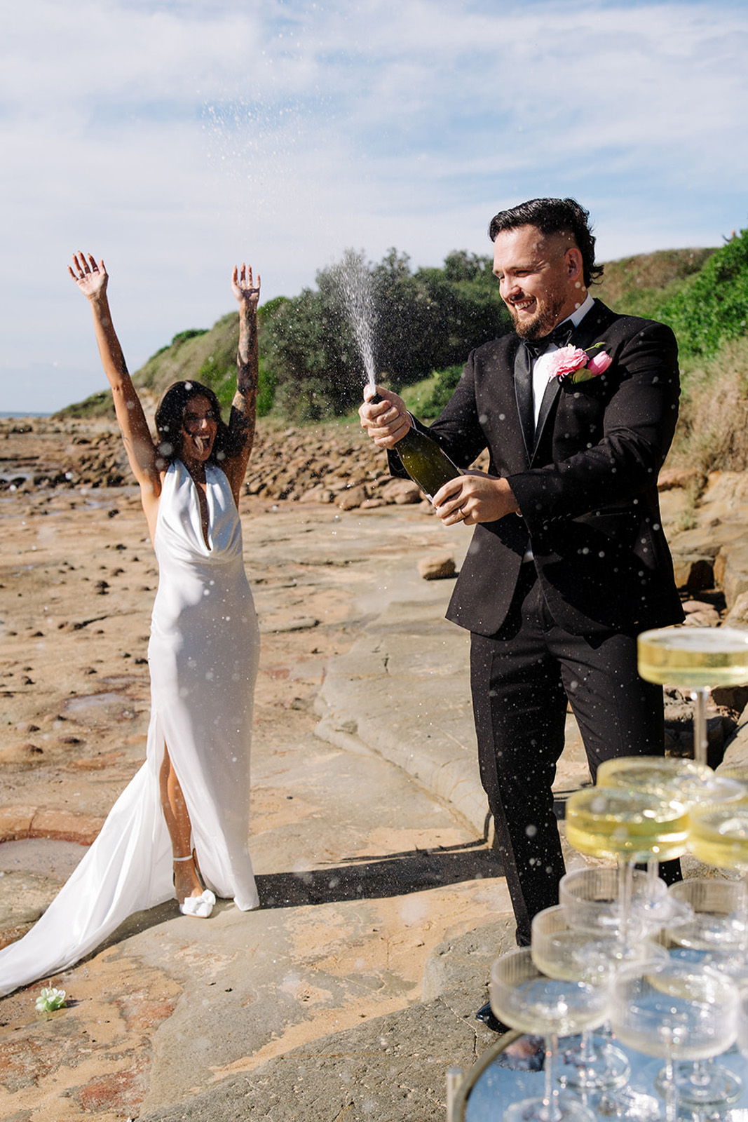 Couple celebrating with champagne on the beach, surrounded by glasses.