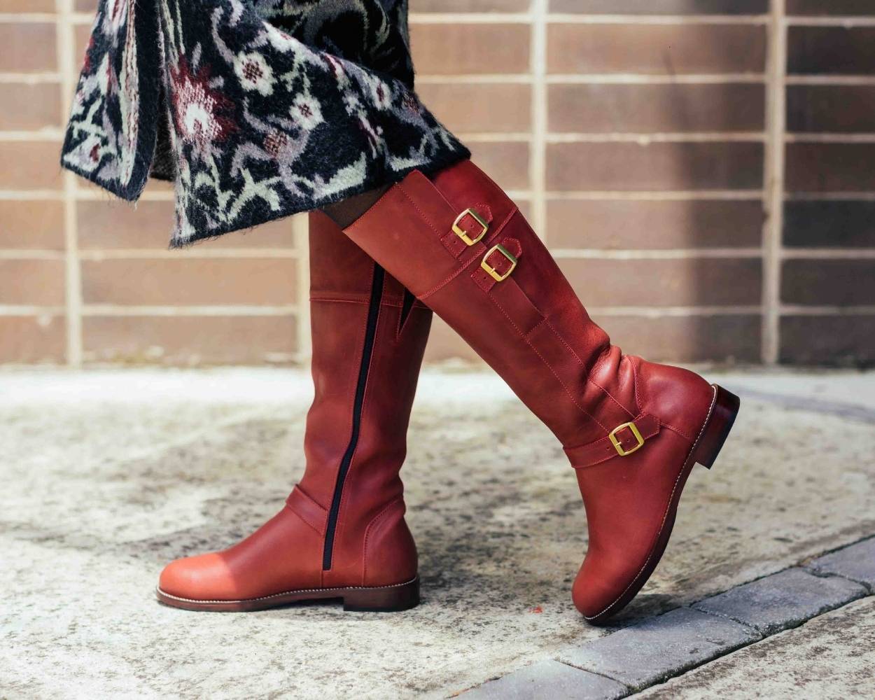 An outdoor lifestyle shot from the knees down of a person walking on a sidewalk next to a brick wall. They are wearing a black patterned coat or skirt and a pair of tall, knee-high red leather boots featuring dual gold buckle straps at the top calf, side zippers, and a low stacked leather heel.