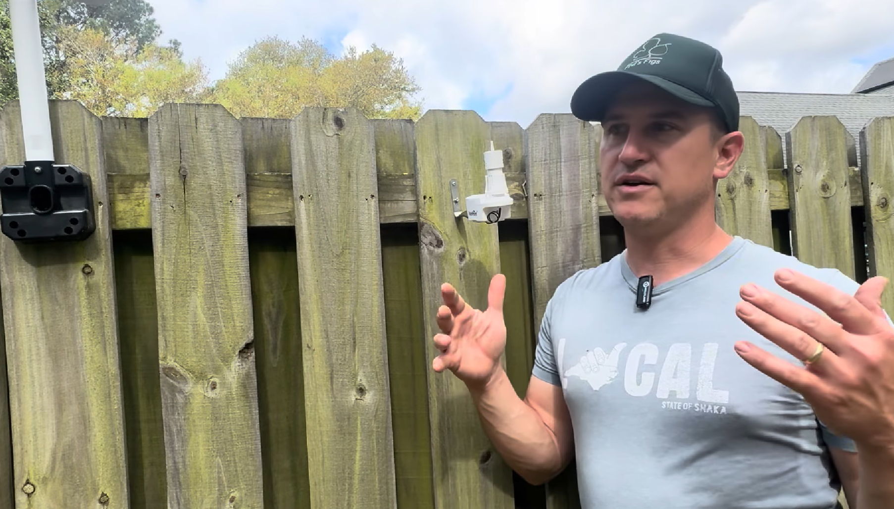 Man gesturing near a wooden fence, wearing a cap and casual shirt.