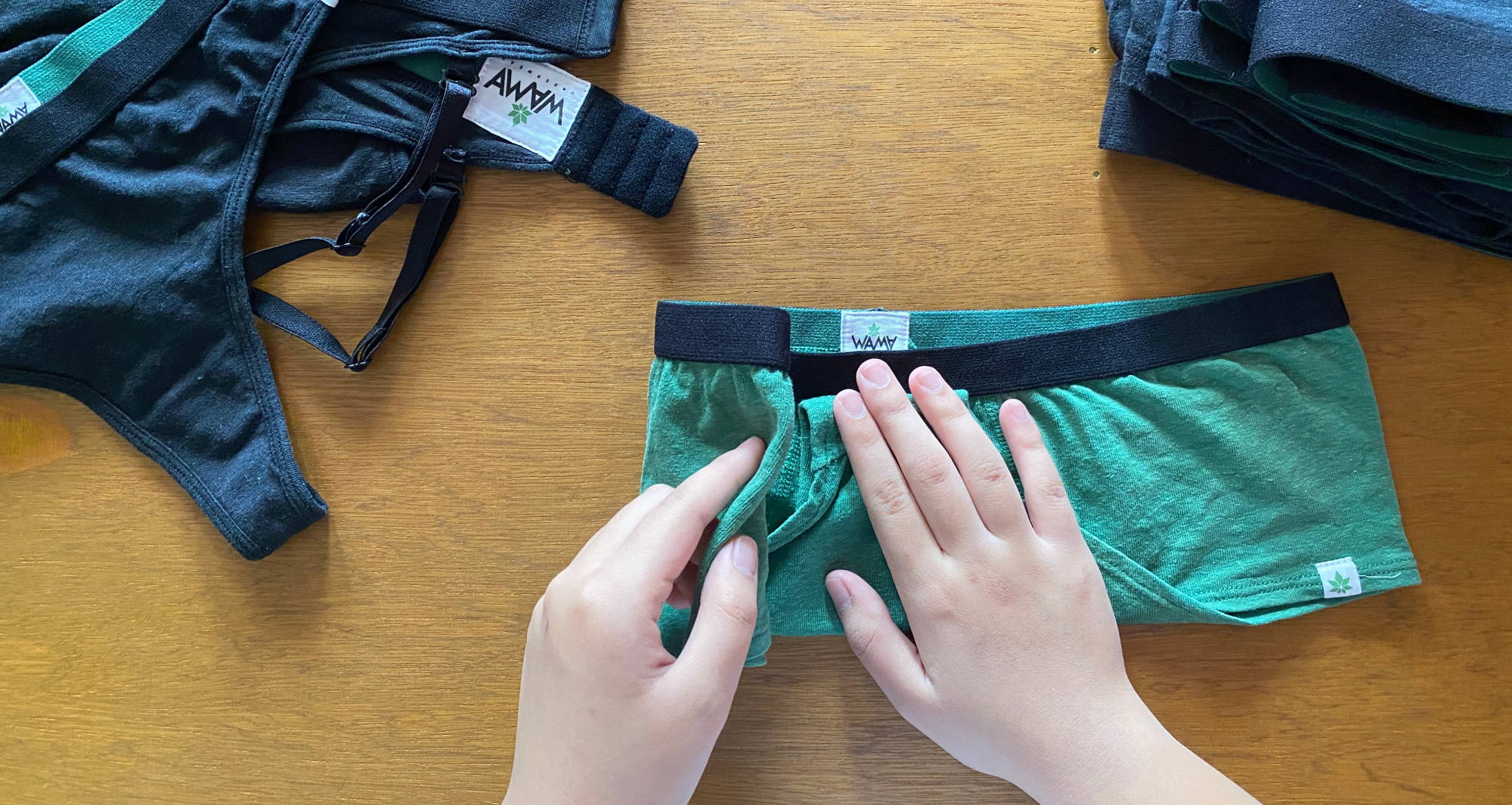 A person’s hands folding a pair of green underwear on a wooden surface next to a pile of unfolded undergarments and a pile of folded underwear