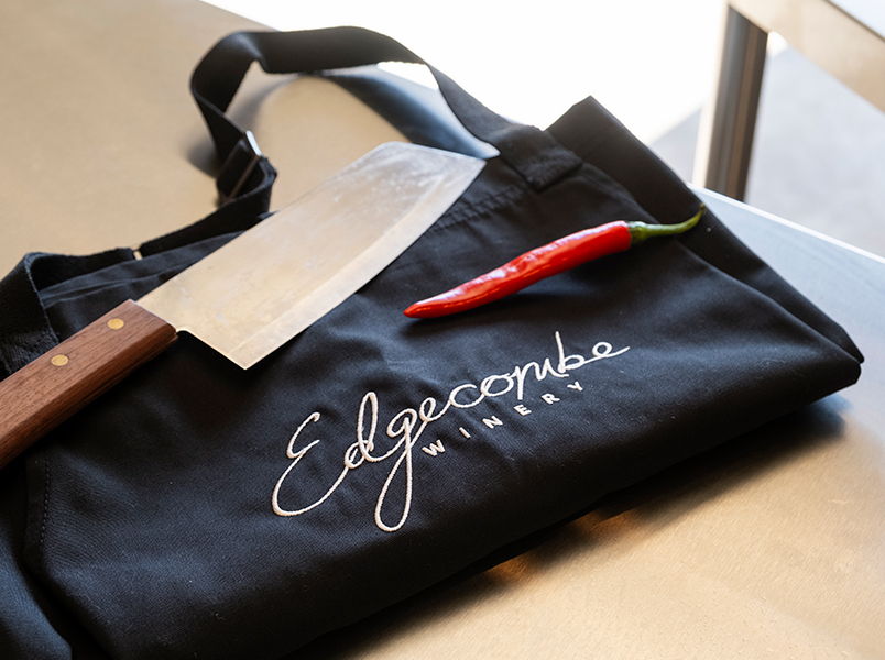 Black folded apron with Edgecombe Winery embroidered in white script on the front, displayed with a knife and chilli pepper sitting on a stainless steel bench